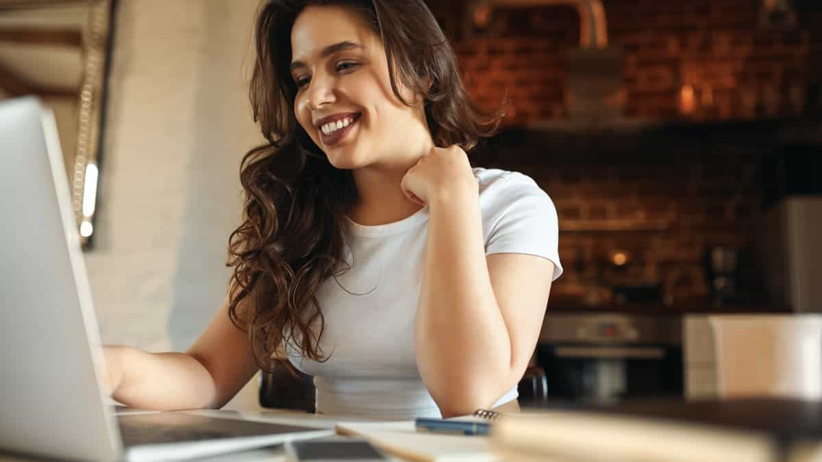 Mulher sorridente utilizando um notebook em um ambiente doméstico aconchegante, transmitindo tranquilidade e foco. A cena remete ao planejamento financeiro e à busca por informações sobre investimentos para aposentadoria, refletindo a importância de preparar o futuro com segurança e autonomia.