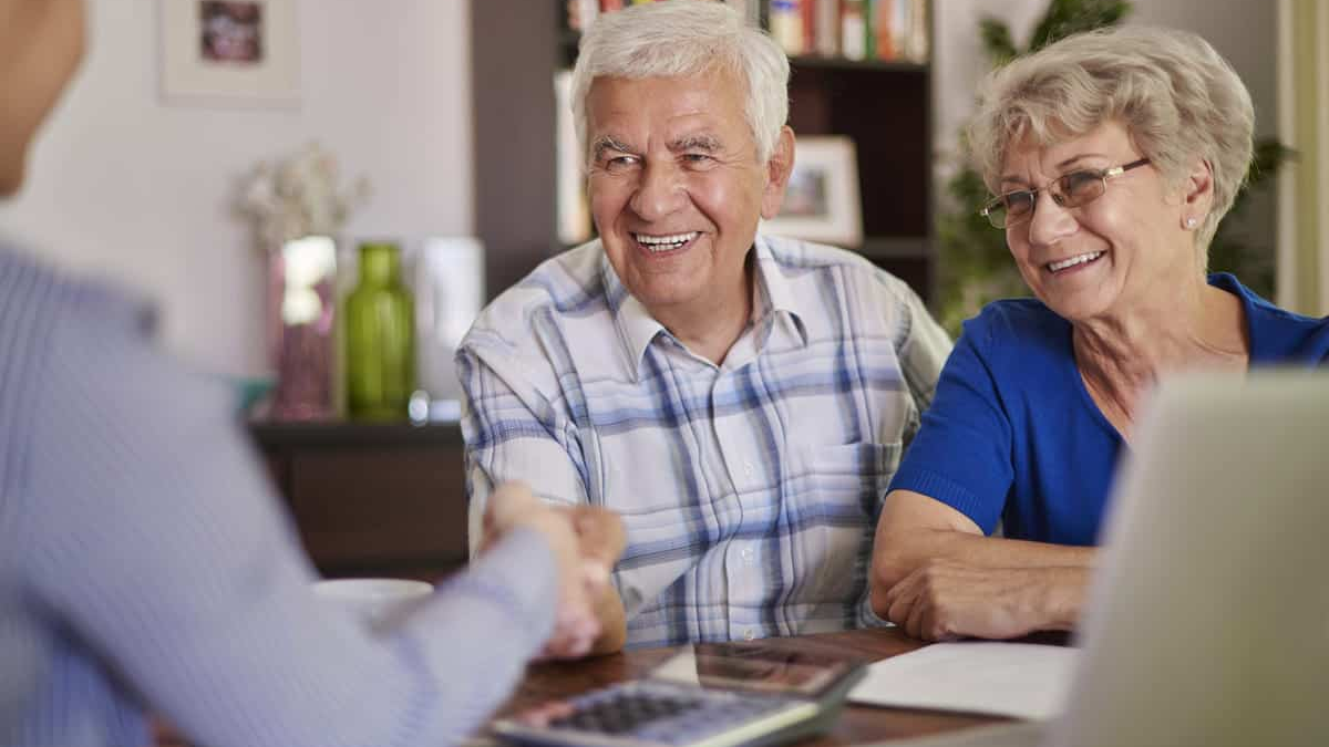 Um casal de idosos sorridente sentado à mesa, conversando com uma terceira pessoa em um ambiente acolhedor. A expressão de tranquilidade e contentamento transmite confiança e bem-estar, refletindo a segurança financeira e a qualidade de vida conquistadas após o planejamento da aposentadoria.