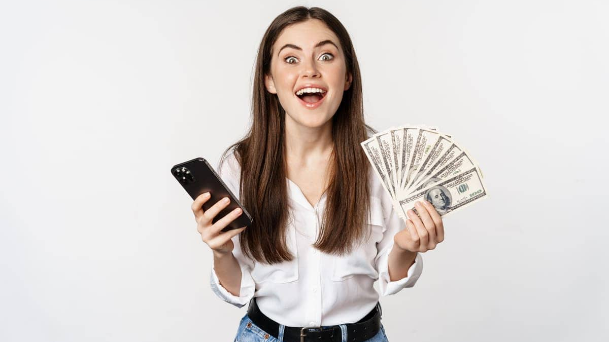 Mulher sorridente segurando um celular em uma mão e várias notas de dinheiro na outra, transmitindo alegria e satisfação. A cena representa o sucesso em conquistar uma renda extra e o entusiasmo de alcançar maior independência financeira.